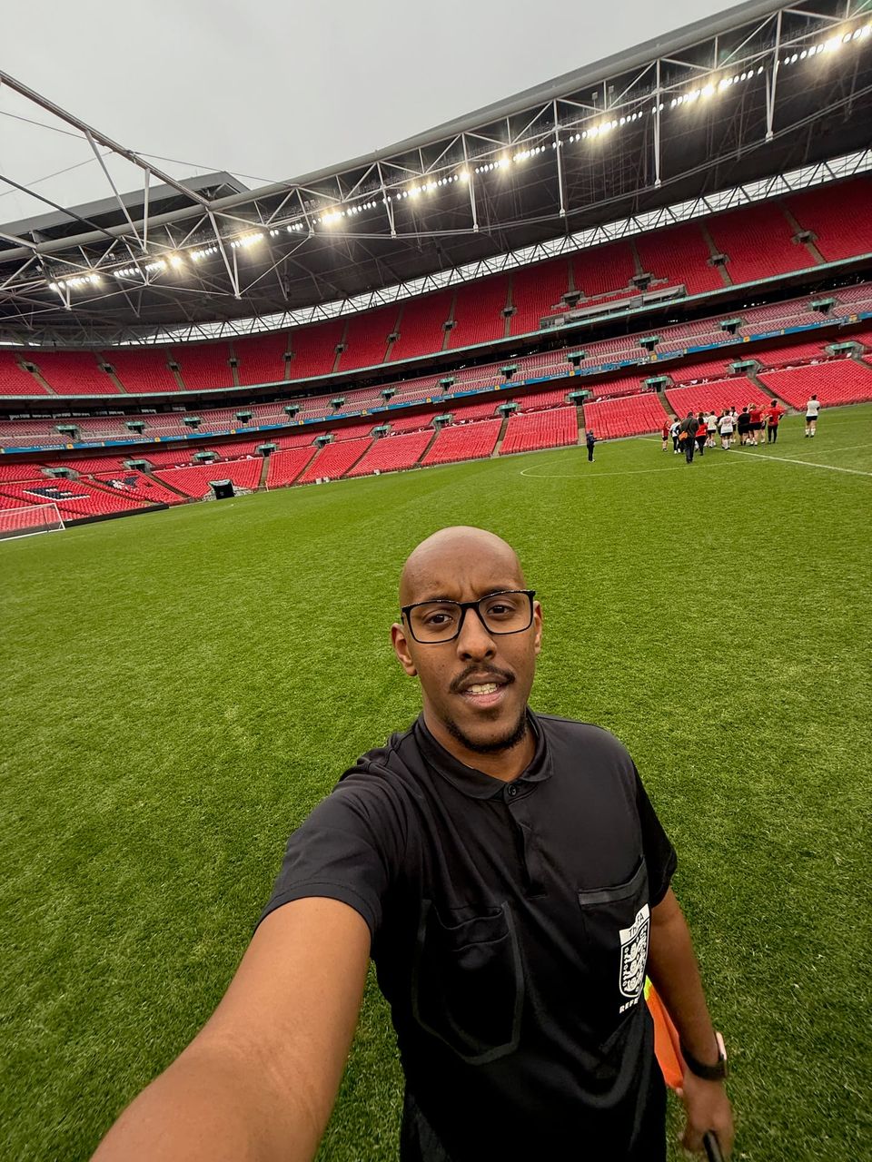 Abdul in a black referee’s kit, holding an assistant referee’s flag on the pitch at Wembley Stadium.
