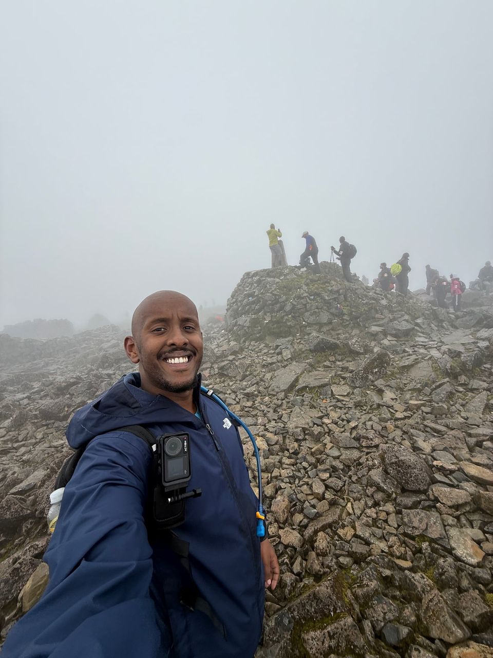 Abdul smiling at a misty, rocky summit, with other walkers gathered at the top behind him.